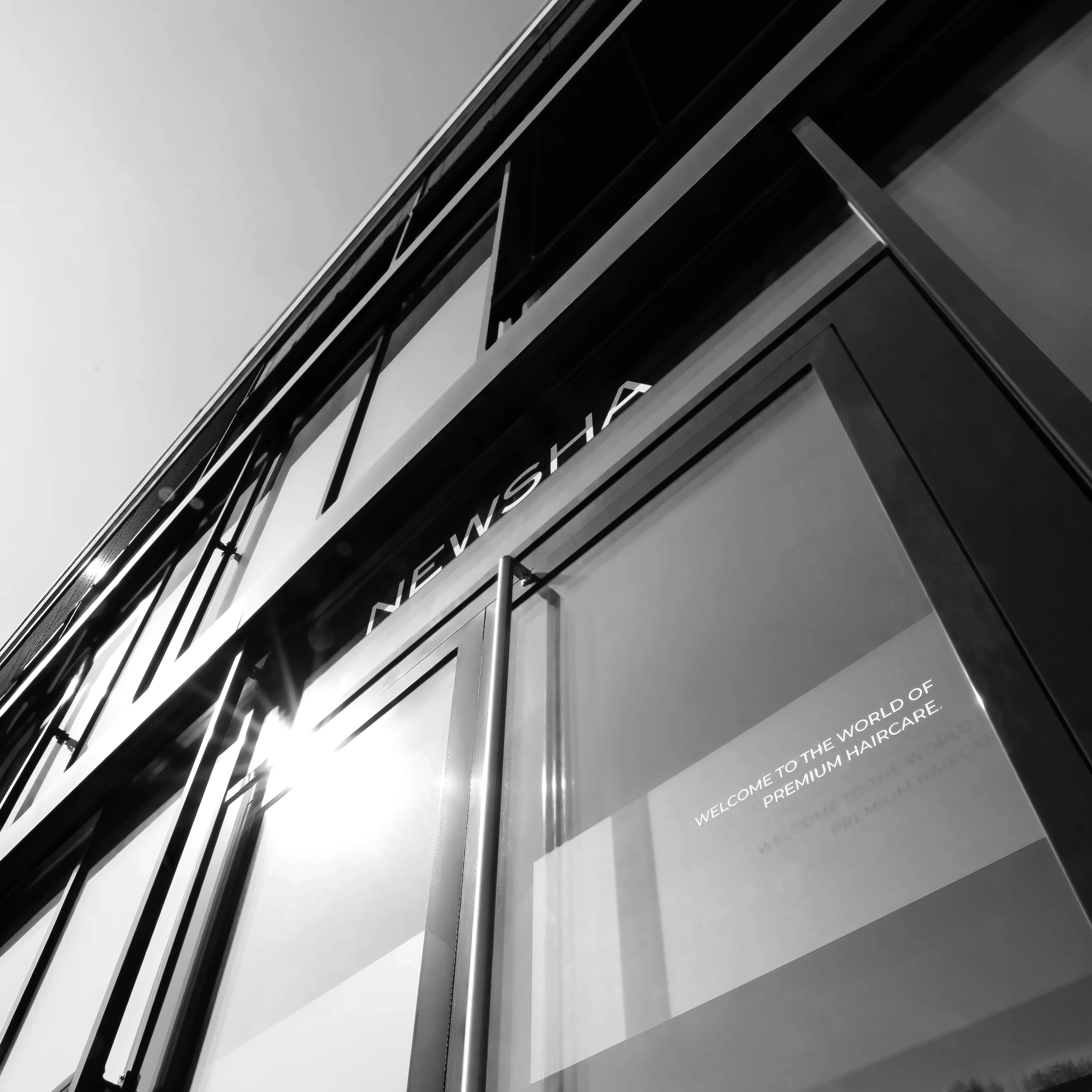 Schwarz-weiße Außenaufnahme des NEWSHA Gebäudes mit moderner Glasfassade, NEWSHA Schriftzug und dem Slogan „Welcome to the World of Premium Haircare“.
