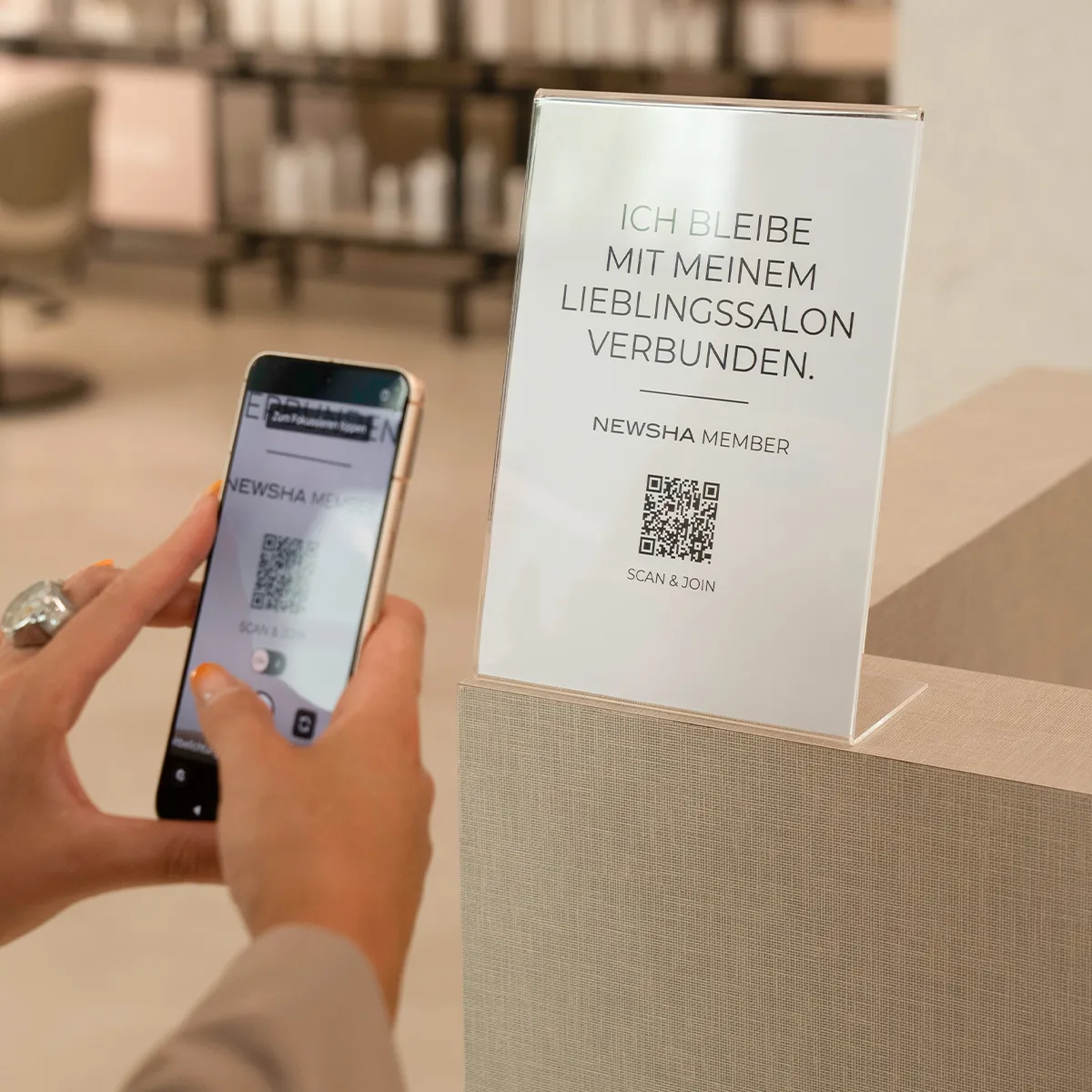 Eine Person scannt mit ihrem Smartphone den QR-Code eines NEWSHA Member Aufstellers im Friseursalon, um dem Kundenprogramm beizutreten.
