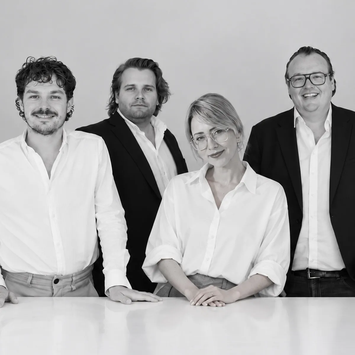 Schwarz-weißes Gruppenporträt des NEWSHA Teams mit drei Männern und einer Frau in eleganter Businesskleidung vor einem hellen Hintergrund.