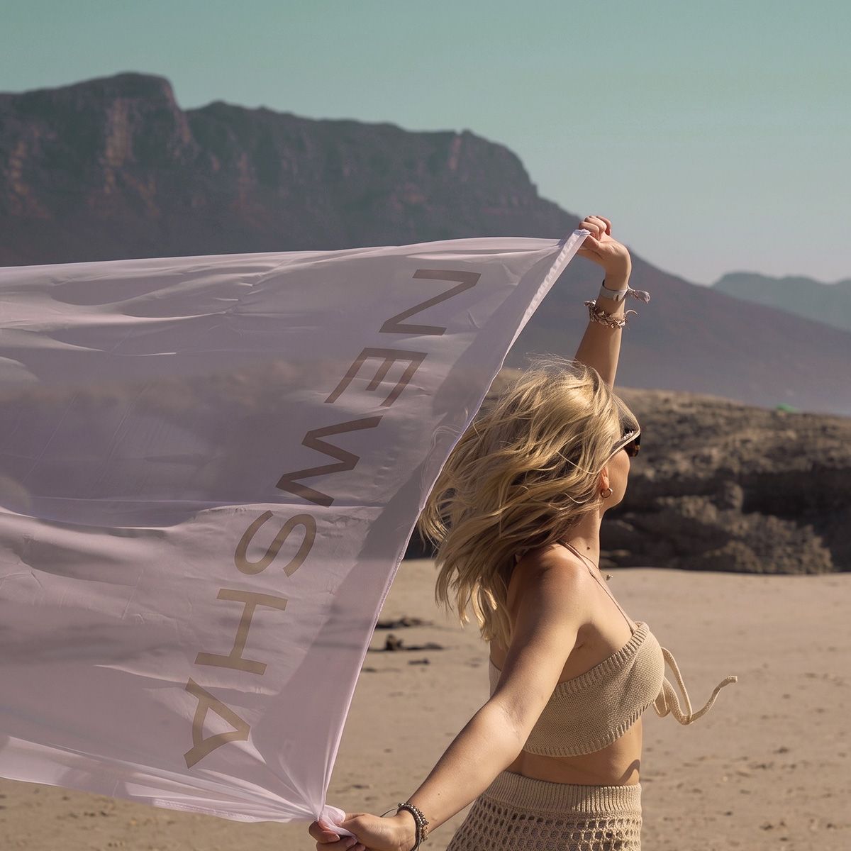 Blonde Frau mit langen, gewellten Haaren h&auml;lt eine gro&szlig;e NEWSHA Flagge in einer sonnigen Landschaft mit Bergen im Hintergrund.