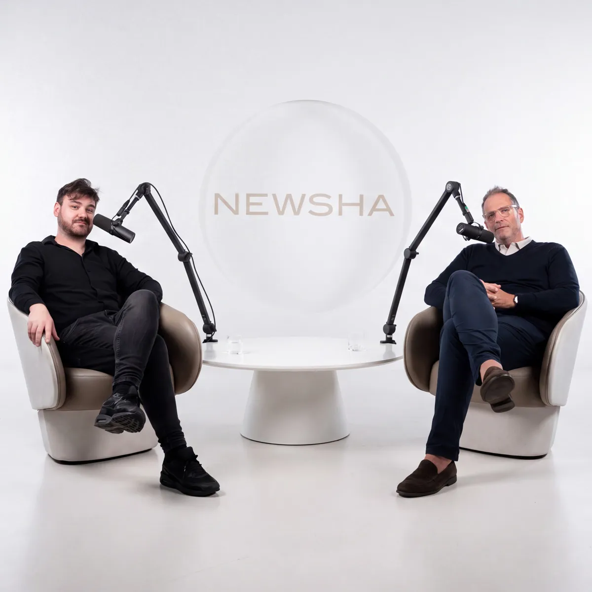 Zwei M&auml;nner sitzen bei einer NEWSHA Podcast-Aufnahme in modernen Sesseln und sprechen vor Mikrofonen mit NEWSHA Schriftzug im Hintergrund.