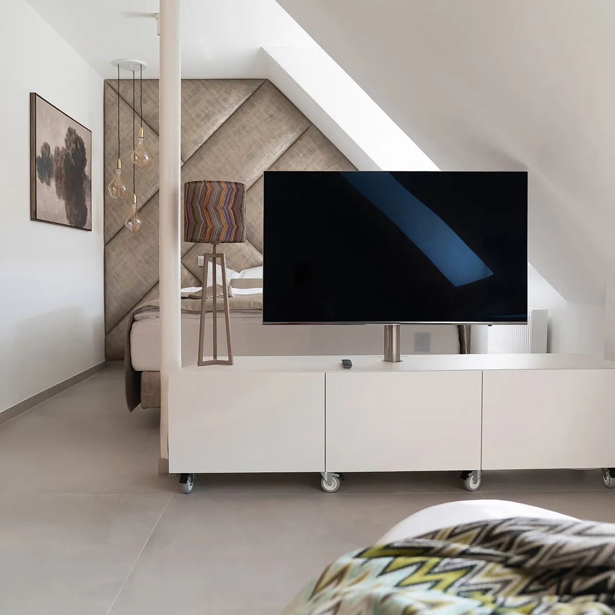 Modernes Apartment unter einer Dachschräge mit Doppelbett, großem Fernseher, weißen Möbeln und stilvoller Beleuchtung.