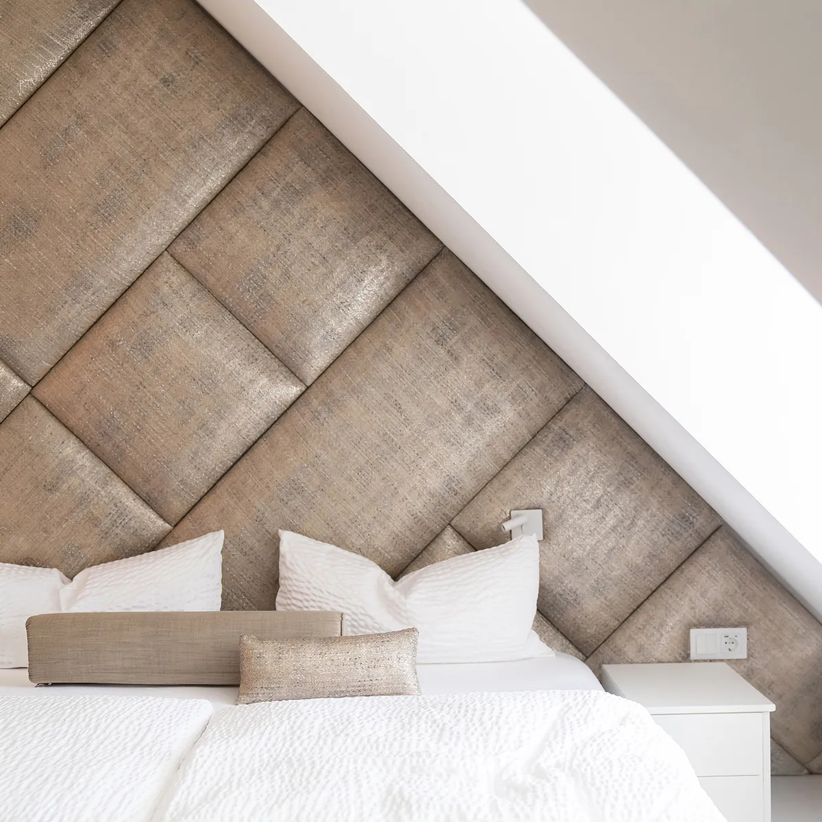 Helles Schlafzimmer mit weiß bezogenem Doppelbett, dekorativen Kissen und einer geometrisch gepolsterten Wand unter einer Dachschräge.