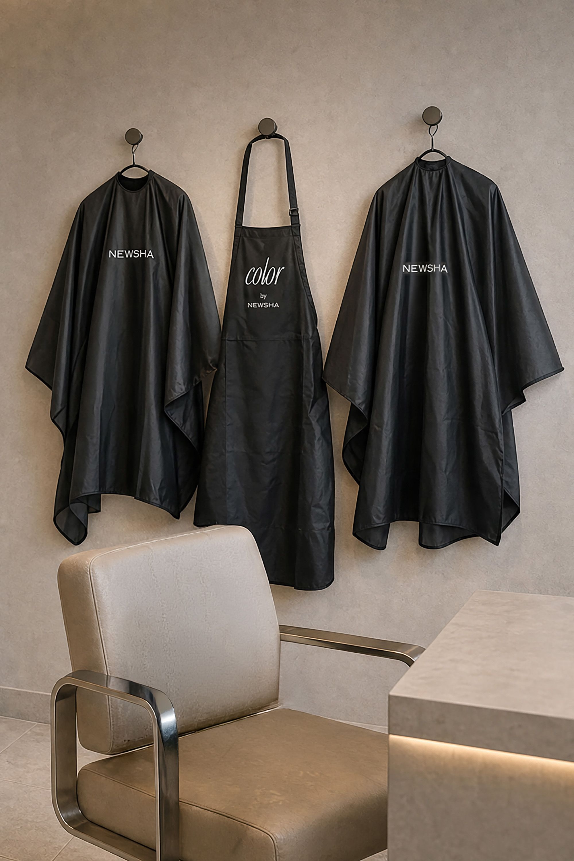Zwei schwarze NEWSHA Friseurumhänge und eine Color by NEWSHA Schürze hängen an einer beigen Salonwand über einem modernen Friseurstuhl.