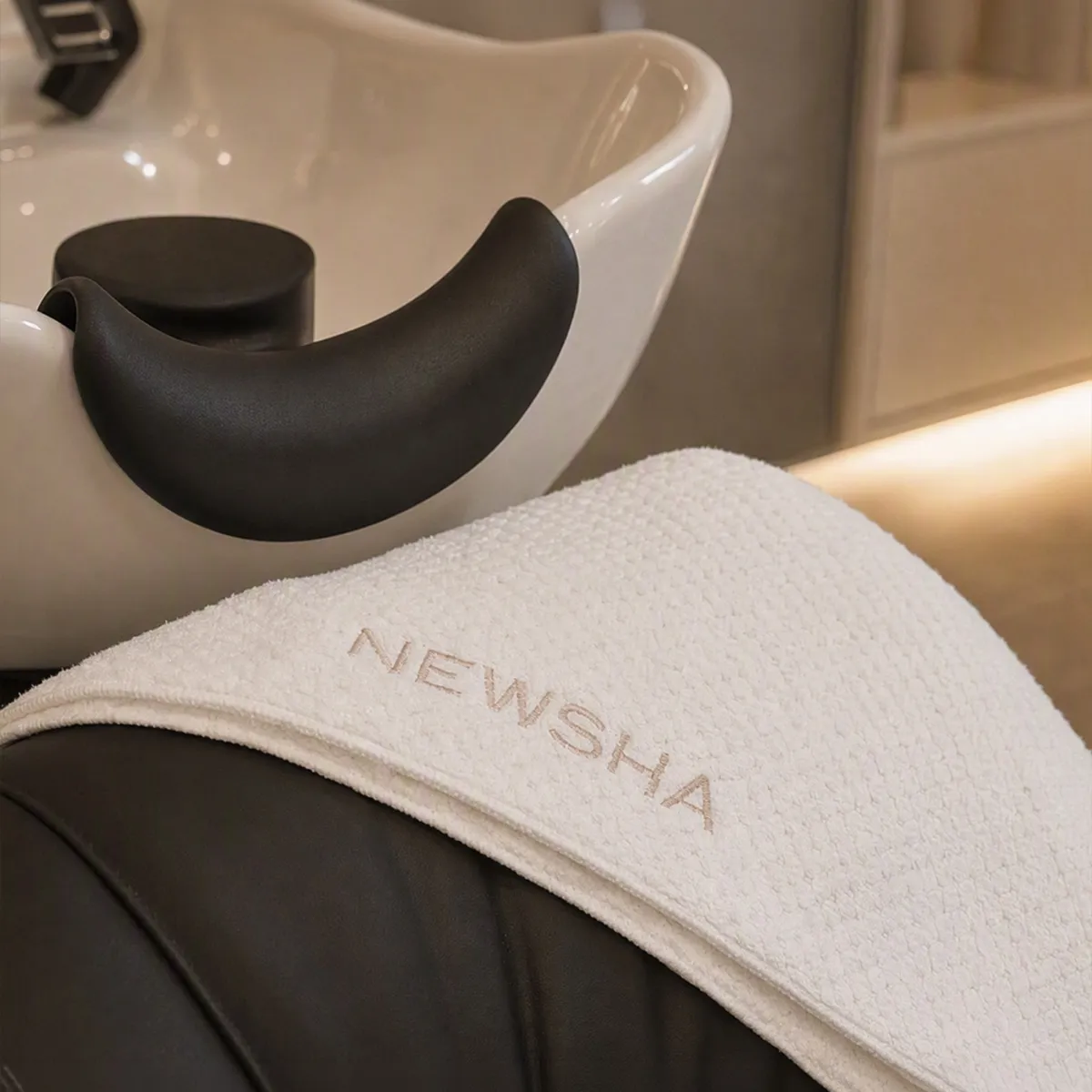 Beiges NEWSHA Handtuch mit gesticktem Markenlogo liegt auf einem schwarzen Salonwaschstuhl neben einem weißen Haarwaschbecken.